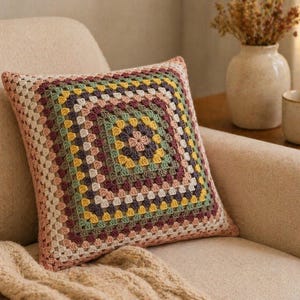 May include: A square throw pillow featuring a multi-colored crochet granny square pattern. The design consists of concentric rows in shades of dusty pink, sage green, mustard yellow, cream, and dark charcoal, creating a textured geometric look.