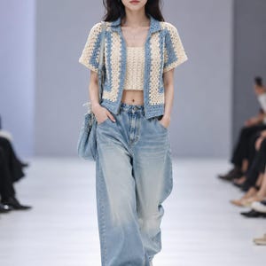 May include: A light blue and cream crochet top and matching short-sleeved jacket paired with light wash, wide-leg jeans. The outfit is accessorized with a light blue shoulder bag and white trainers. The model is walking on a white runway.