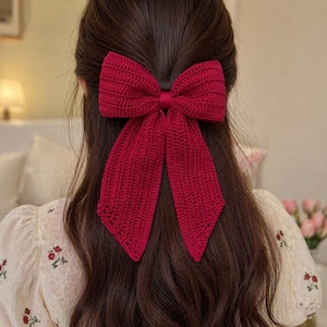 以下が含まれることがあります： 大きな赤いニットのヘアクリップが後頭部に取り付けられています。 弓は質感のある素材でできており、長く流れるようなテールが付いています。 髪は長く、濃い茶色です。 その人は、花の刺繍が施されたクリーム色のトップスを着ています。