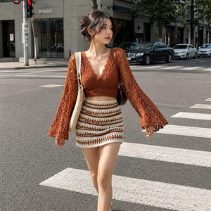 Puede incluir: Un top corto de ganchillo color óxido de manga larga con escote en V y mangas acampanadas, combinado con una minifalda de ganchillo a rayas horizontales en crema, marrón y óxido. Un bolso de mano tejido en color beige se lleva al hombro.