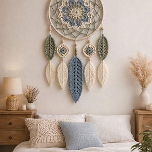 Op de afbeelding: Een handgemaakte dromenvanger met een crèmekleurig cirkelvormig frame en een centraal blauw en crèmekleurig bloemmotief. Het heeft hangende gehaakte veren en bladvormen in crème en blauw, wat een bohemien esthetiek creëert. De dromenvanger wordt boven een bed getoond.