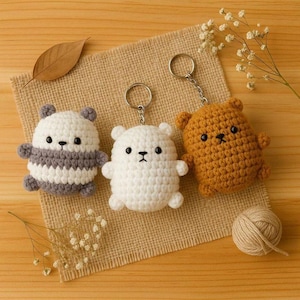 Pode incluir: Três chaveiros de urso de crochê feitos à mão em branco, marrom e panda em preto e branco. Cada urso tem um pequeno chaveiro de metal anexado. Os ursos são exibidos em um pano de juta com flores secas e uma bola de lã.
