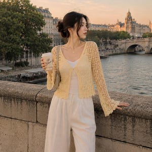 May include: A pale yellow long-sleeve crochet cardigan featuring a scalloped hem and bell sleeves, worn over a white camisole and paired with white wide-leg trousers. The cardigan has a delicate open-knit pattern and a single front button closure.