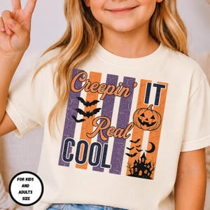 Puede incluir: Una camiseta color crema con un gráfico desgastado de rayas verticales moradas y naranjas. El texto "Creepin' IT Real COOL" aparece junto a ilustraciones de murciélagos, una calabaza, una luna creciente y la silueta de una casa encantada.