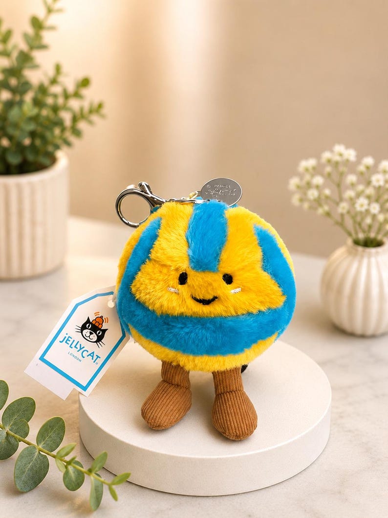 May include: A plush, round keychain toy with a smiling face, featuring yellow and blue stripes. It has brown corduroy legs and a silver keyring. A tag with the text "JELLYCAT LONDON" is attached.