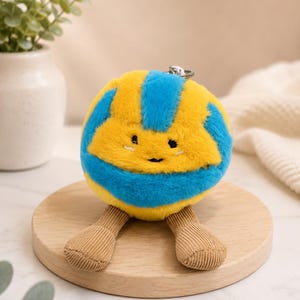 May include: A plush yellow and blue ball-shaped keychain with a smiling face and corduroy legs. The keychain is on a wooden round base, with a silver metal ring attached to the top.