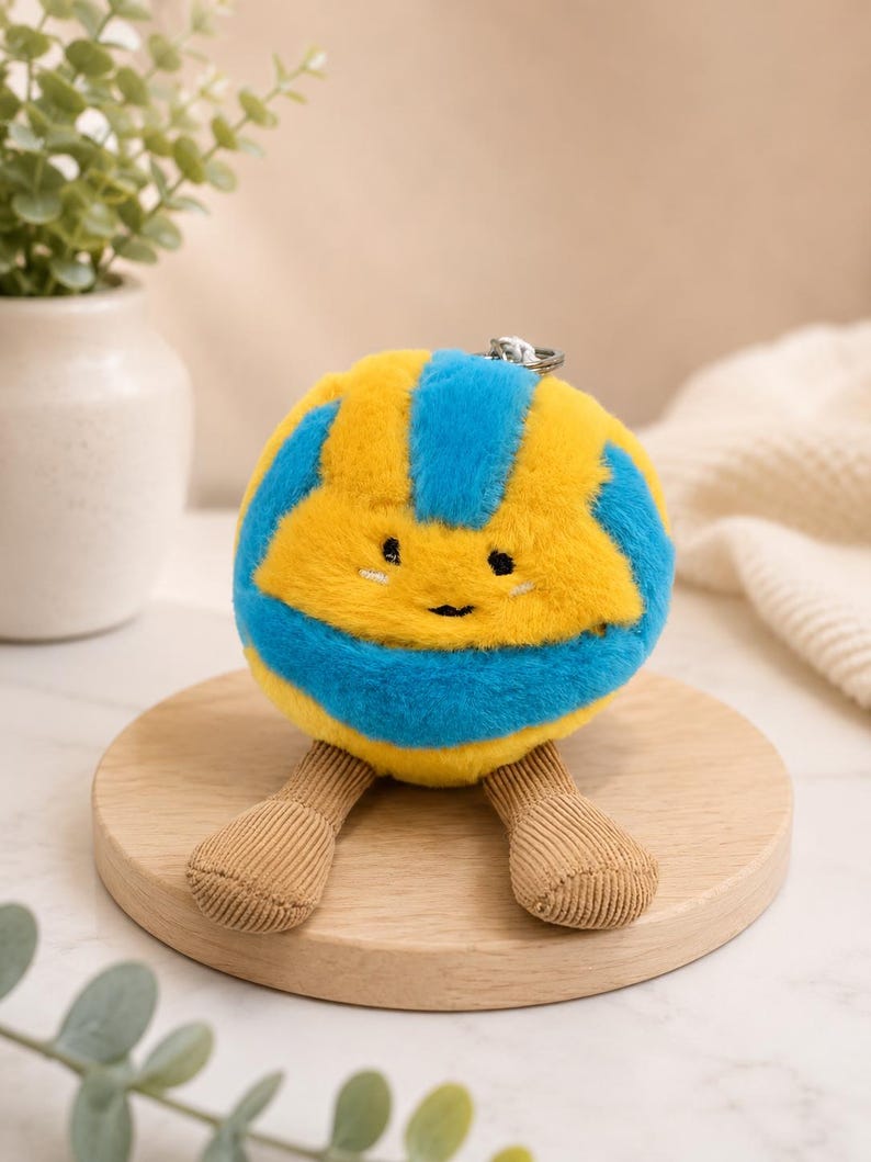 May include: A plush yellow and blue ball-shaped keychain with a smiling face and corduroy legs. The keychain is on a wooden round base, with a silver metal ring attached to the top.