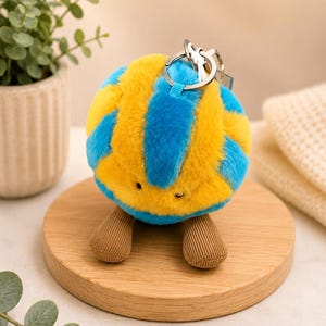 May include: A plush, spherical keychain with a blue and yellow striped pattern. The keychain has small, brown, corduroy-like feet and a silver keyring. The item is displayed on a wooden coaster, with greenery in the background.