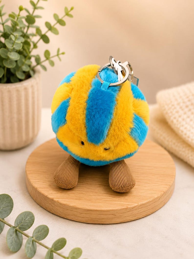 May include: A plush, spherical keychain with a blue and yellow striped pattern. The keychain has small, brown, corduroy-like feet and a silver keyring. The item is displayed on a wooden coaster, with greenery in the background.