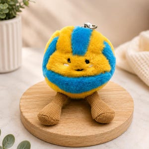 May include: A plush toy in the shape of a smiling ball, featuring yellow and blue stripes. It has small brown corduroy legs and a silver keychain. The toy is placed on a round wooden stand.