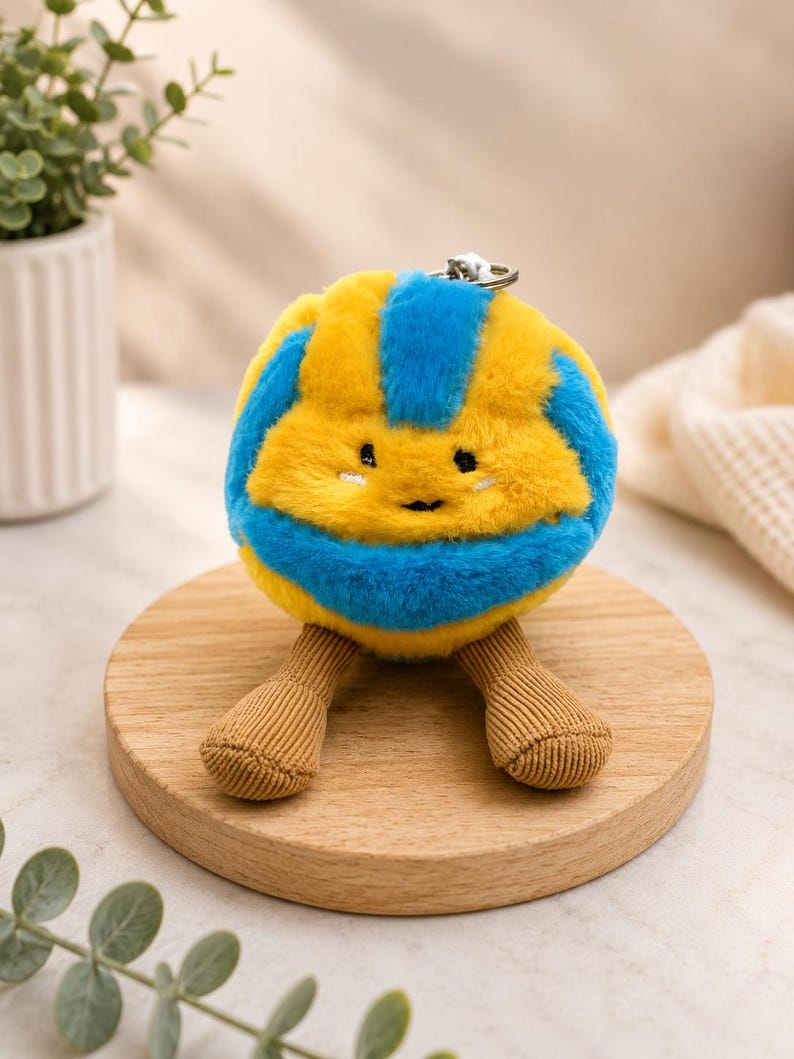 May include: A plush toy in the shape of a smiling ball, featuring yellow and blue stripes. It has small brown corduroy legs and a silver keychain. The toy is placed on a round wooden stand.
