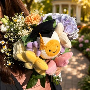 May include: A colorful bouquet featuring a plush egg character wearing a graduation cap. The bouquet includes yellow, pink, and purple plush flowers, and real flowers in shades of white, orange, and purple. The background is a blurred outdoor scene.