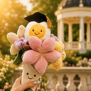 May include: A plush bouquet featuring a smiling fried egg wearing a graduation cap, surrounded by fabric flowers in shades of pink, yellow, and purple. The bouquet is wrapped in a cone with a smiling face.
