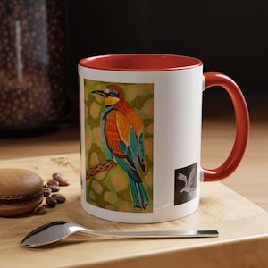 Könnte beinhalten: Weißer Keramikbecher mit roter Innenseite und Henkel. Der Becher zeigt eine farbenfrohe Illustration eines Vogels, der auf einem Ast sitzt. Ein kleines schwarzes Quadrat mit einer weißen Vogelgrafik befindet sich an der Seite.