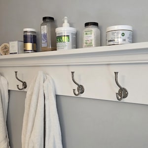May include: A white wall-mounted coat rack with a top shelf featuring crown molding. The rack includes three double-pronged metal hooks in a brushed finish. Two white towels hang from the hooks, and various toiletry containers sit on the shelf.