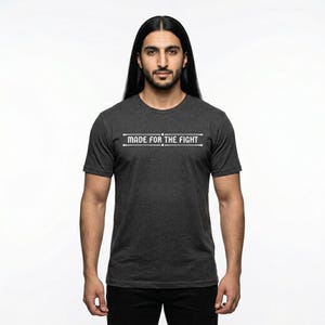 May include: Dark grey t-shirt with the white text "MADE FOR THE FIGHT". The short-sleeved shirt has a crew neck. The person in the image is wearing black trousers.
