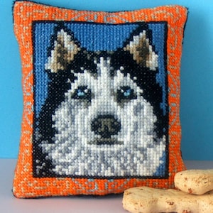 May include: A small orange and blue cushion with a cross-stitch image of a husky dog's face. The dog has blue eyes and a white and black coat.