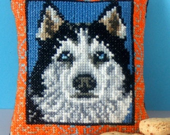 May include: A small orange and blue pillow with a cross-stitch image of a husky dog's face. The dog has blue eyes and a white and black coat.