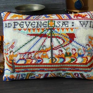 May include: A rectangular cushion with a detailed cross-stitch design. The design features a boat with figures, using red, blue, yellow, and green threads. The text "AD PEVENE...WIL" is stitched across the top. The cushion has a blue border.
