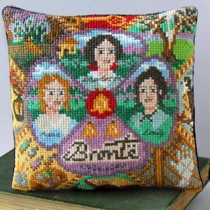May include: A square decorative cushion featuring a colourful cross-stitch design of three women with the names Charlotte, Anne, and Emily stitched below them. The centre displays a red bell icon above the text Brontë in a dark, stylised font.