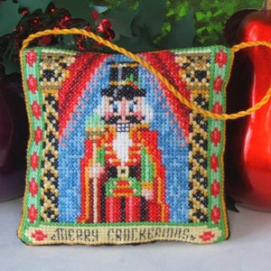 以下が含まれることがあります： 赤、緑、青のデザインのくるみ割り人形が描かれたクロスステッチのクリスマスオーナメント。吊り下げ用の金色のコードと「MERRY CRACKERMAS」の文字があります。背景には赤と緑のホリデーデコレーションがあります。
