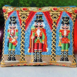 以下が含まれることがあります： 3体のくるみ割り人形のクロスステッチデザインが施された装飾用クッション。人形は青い背景に赤、緑、金のユニフォームを着ています。クッションはベージュの縁取りで、赤と金のアクセントが施されています。
