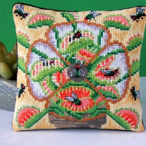 May include: A square pillow with a colorful embroidered design of a carnivorous plant with red, green, and yellow leaves and multiple flies. The pillow is on a white surface.