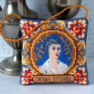May include: A small, square, embroidered pillow with a gold frame and a portrait of a woman with the text "Sherry Fitzroy" at the bottom.