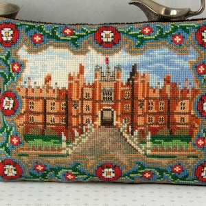 May include: A needlepoint pillow with a colorful embroidered image of a brick building with a large arched entrance. The building is surrounded by a green lawn and a blue sky. The pillow has a red, white, and green border with a floral design.