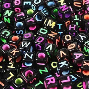 Könnte beinhalten: Nahaufnahme eines Haufens schwarzer Alphabetperlen. Jeder würfelförmige Stein zeigt einen anderen Buchstaben in einer lebendigen, mehrfarbigen Schrift. Die Perlen sind verstreut und bilden eine optisch ansprechende und spielerische Anordnung, ideal für Bastel- oder Bildungszwecke.