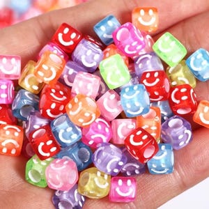 May include: A handful of colorful, cube-shaped beads. Each bead is a different translucent color, including red, blue, pink, and green. A white smiley face is printed on each side of the beads. These beads are ideal for jewelry making and crafts.