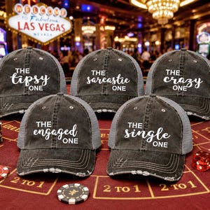 May include: Five black trucker caps with white text: "The Tipsy One," "The Sarcastic One," "The Crazy One," "The Engaged One," and "The Single One." The caps have a grey mesh back. The hats are on a red surface.
