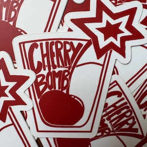 May include: A collection of die-cut stickers featuring a red and white design. Each sticker displays a glass containing a cherry with the text CHERRY BOMB printed above it, accompanied by a separate red and white star-shaped sticker design.