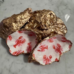 May include: Two decorative oyster shells with a metallic gold exterior finish. The interior of each shell is painted white and features a repeating pattern of small, detailed red lobsters. The shells have irregular, natural edges and textures.