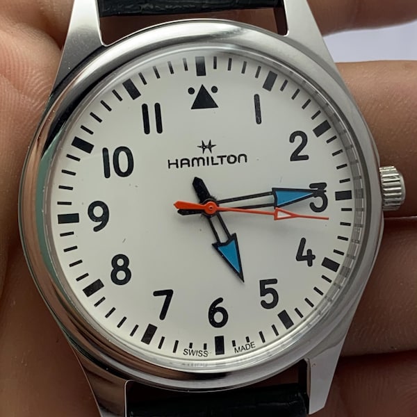 May include: A Hamilton wristwatch with a white dial, black Arabic numerals, and a triangle marker at the twelve o'clock position. The watch features blue-outlined hour and minute hands, a thin orange second hand, and a black leather strap.