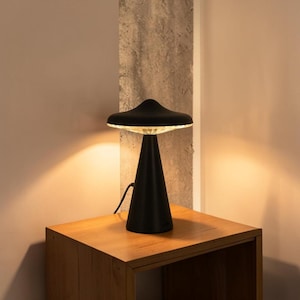 Puede incluir: Una lámpara de mesa negra mate con base cónica y una pantalla ancha en forma de hongo. La parte inferior de la pantalla presenta un difusor texturizado de vidrio o plástico translúcido que emite un brillo cálido y suave hacia abajo.
