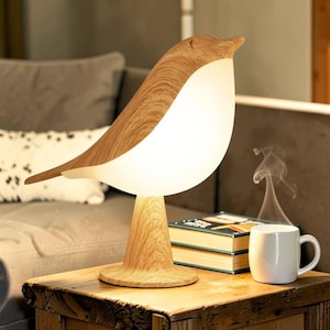 Peut inclure: Lampe de table en forme d'oiseau en bois, avec un corps blanc lumineux et une tête et des ailes en bois brun clair. La lampe est posée sur un socle en bois. Une tasse blanche et des livres sont sur la table en bois.