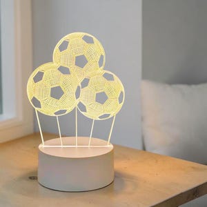 May include: A 3D illusion lamp featuring three illuminated soccer balls arranged in a cluster. The design is composed of thin, glowing white lines that form the hexagonal and pentagonal patterns of the balls, mounted on a solid cylindrical base.