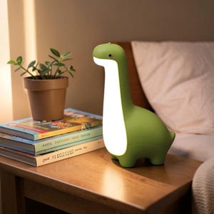May include: A green dinosaur-shaped lamp with a smooth, matte finish and a glowing white belly. The lamp features a long neck, a small head with a single visible eye, and four short legs, emitting a soft, warm light from its front section.