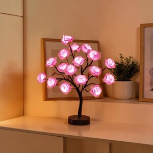 Könnte beinhalten: Eine dekorative baumförmige Lampe mit rosa rosenförmigen Lichtern. Die Lampe hat einen schwarzen Sockel und Stiel, mit mehreren Zweigen, die die beleuchteten Rosen halten. Der Hintergrund umfasst ein gerahmtes Bild und eine kleine Topfpflanze.