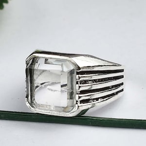 May include: A silver ring with a clear, rectangular gemstone setting. The band has etched horizontal lines. The ring is displayed against a white background, accentuating its design. The ring's design is simple and elegant.