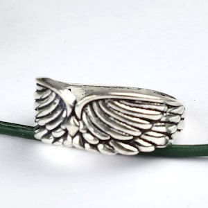May include: A silver-toned ring featuring a detailed, symmetrical wing design that wraps around the band. The metal has a polished finish with darkened recessed areas to highlight the individual feather textures carved into the surface.