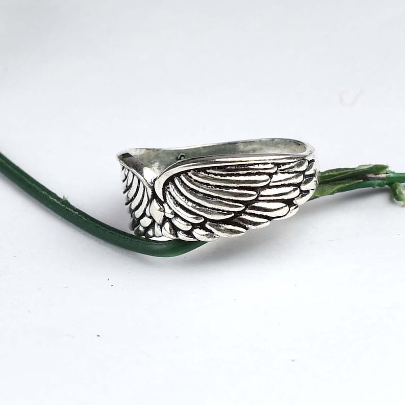 May include: A silver-toned metal ring featuring a detailed, embossed wing design that wraps around the band. The surface of the ring has a polished finish with darkened recessed areas to highlight the individual feather textures of the wing motif.