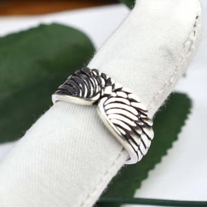 May include: A silver-toned metal ring featuring a detailed design of two symmetrical feathered wings that meet at the center. The surface of the wings has oxidized dark recessed areas to highlight the individual feather textures and shapes.
