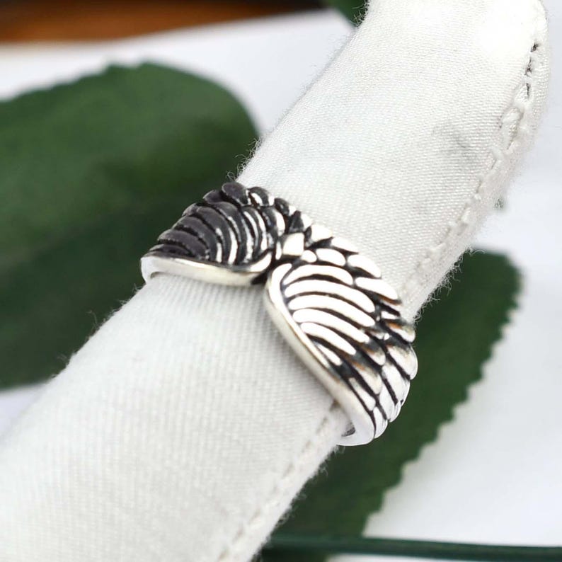 May include: A silver-toned metal ring featuring a detailed design of two symmetrical feathered wings that meet at the center. The surface of the wings has oxidized dark recessed areas to highlight the individual feather textures and shapes.
