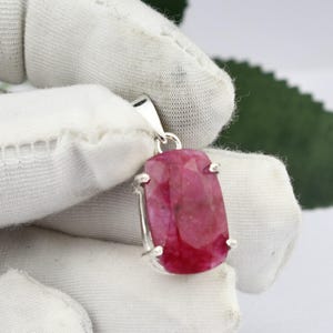 Peut inclure: Un pendentif en rubis facetté de forme rectangulaire monté sur une monture en métal argenté avec une bélière sur le dessus. La pierre précieuse présente une teinte rouge rosée profonde avec des inclusions naturelles et une surface polie.