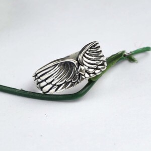 May include: A silver-toned metal ring featuring a detailed, sculpted wing design that wraps around the band. The surface of the wings displays deep, dark oxidized grooves to emphasize the individual feather textures and the overall shape.