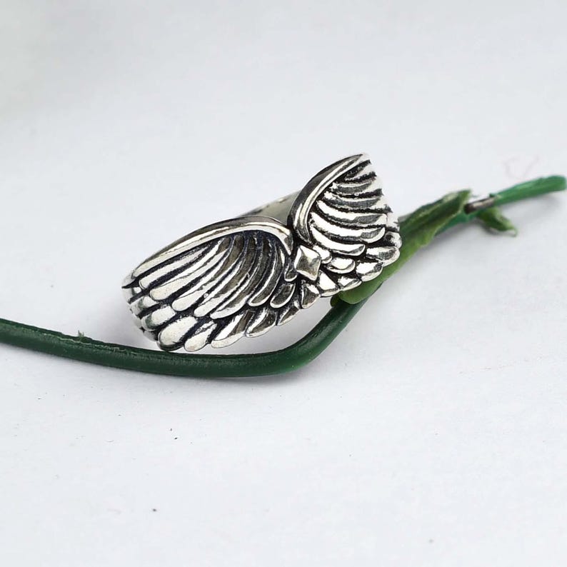May include: A silver-toned metal ring featuring a detailed, sculpted wing design that wraps around the band. The surface of the wings displays deep, dark oxidized grooves to emphasize the individual feather textures and the overall shape.