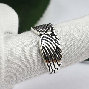 May include: A silver-toned metal ring featuring a detailed, raised wing design that wraps around the band. The surface of the ring has darkened, recessed areas to emphasize the individual feather textures and the overall shape of the wings.