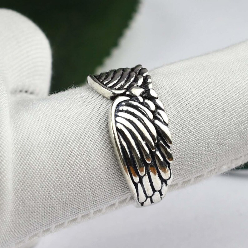 May include: A silver-toned metal ring featuring a detailed, raised wing design that wraps around the band. The surface of the ring has darkened, recessed areas to emphasize the individual feather textures and the overall shape of the wings.
